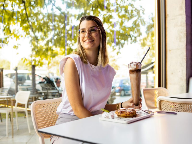 A young and pretty woman with glasses stands inside a modern cafe having a snack.Beautiful woman drinks chocolate milkshake and strawberry and cream pancakes. Concept of women eating sweets. A young and pretty woman with glasses stands inside a modern cafe having a snack.Beautiful woman drinks chocolate milkshake and strawberry and cream pancakes. Concept of women eating sweets.